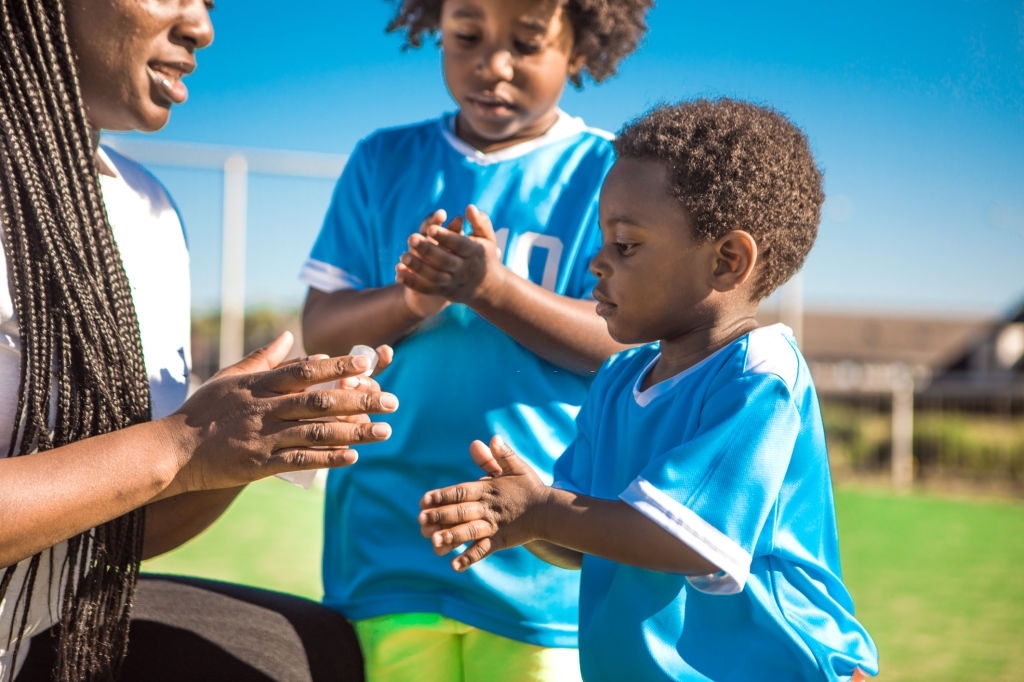 Mother young sons cleaning their hands with gel whilst playing soccer on a football pitch on a beautiful sunny day